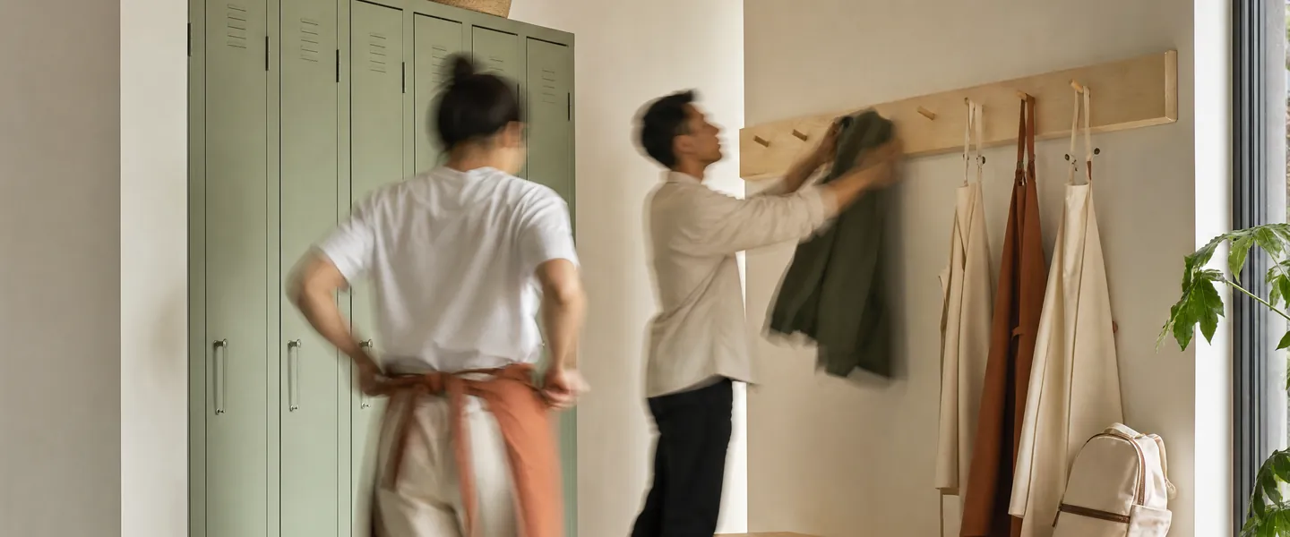 Dos personas del equipo en el vestuario de un local, al empezar el turno: una se ata el delantal y otra cuelga su chaqueta junto a los lockers.