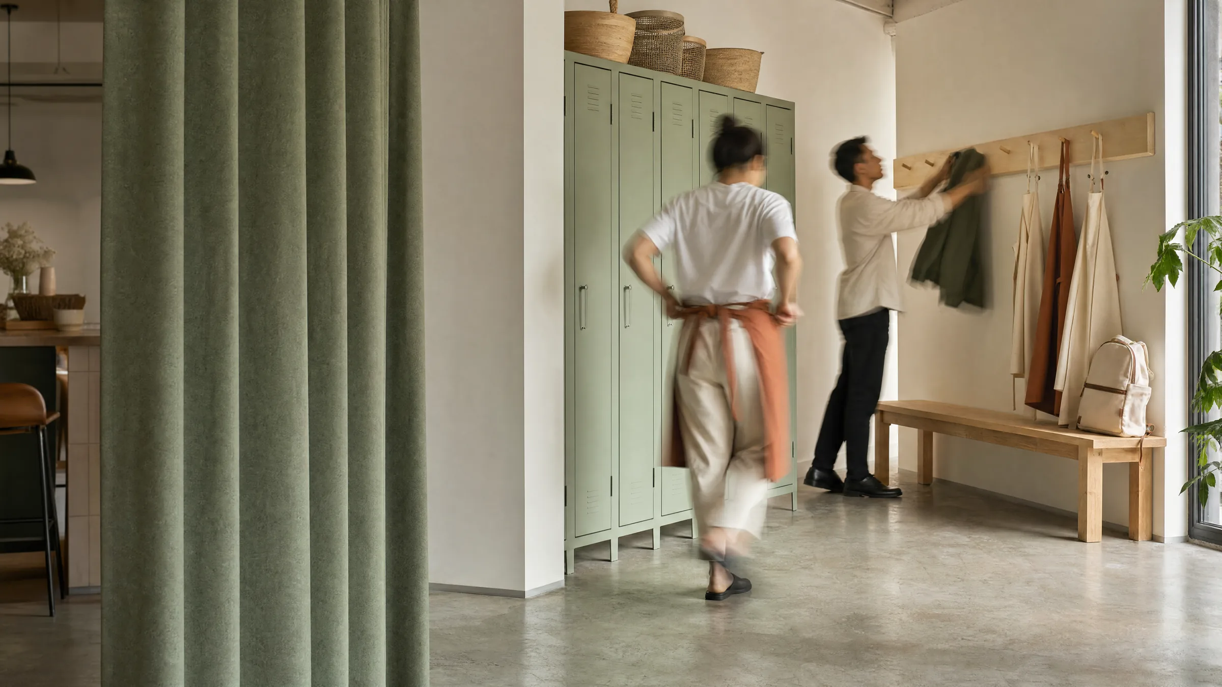 Dos personas del equipo en el vestuario de un local, al empezar el turno: una se ata el delantal y otra cuelga su chaqueta junto a los lockers.